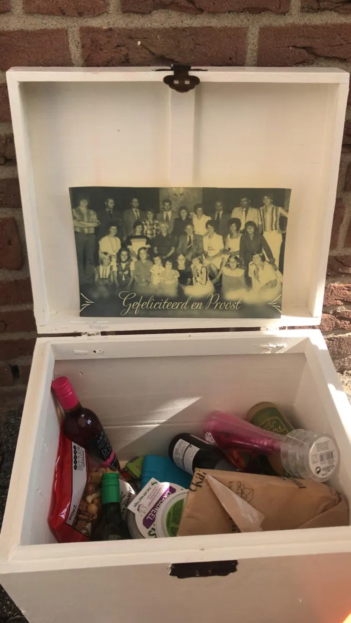Een open witte kist met een oude familiefoto in het deksel, gevuld met kaas, drankjes en hapjes